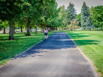 A person walks along an asphalt pathway running through a lush green grass area surrounded by trees